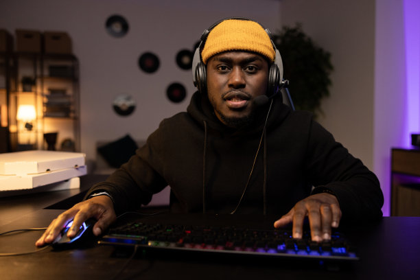 一名戴着黄色帽子戴着耳机的严肃男子正在电脑上认真地玩视频射击游戏。在线多人游戏锦标赛。背景是一个夜晚漆黑的房间，里面堆满了披萨盒图片下载