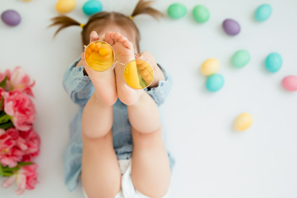 一个孩子躺在有复活节彩蛋的白色背景上，玩着黄色的眼镜。图片下载