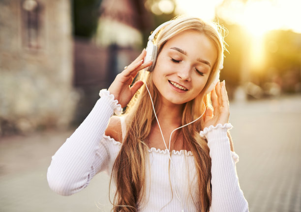 在一个阳光明媚的日子里，一个十几岁的女孩享受着生活，走在街上，头戴耳机听着音乐，以落日为背景图片下载
