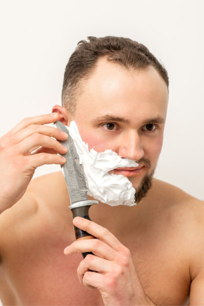 年轻的白人男子剃胡子用一把大刀在白色的背景。图片下载