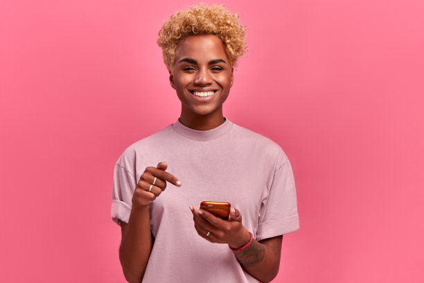 快乐的非裔美国人女士大大的微笑，指着手机屏幕，为有了新的应用程序而高兴，享受在线交流，情绪高昂，在粉红色的背景下孤立。技术图片下载