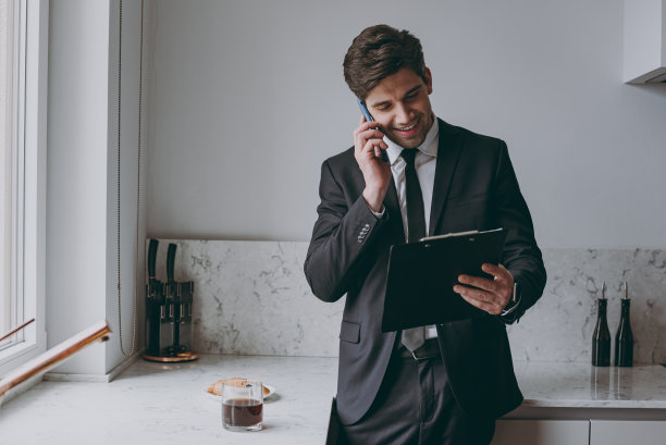 年轻的雇员，商人，公司律师，穿着正式的黑色西装，衬衫，领带，说话，手机，拿着纸夹，文件，站在厨房，在家，成就职业生活方式的概念图片下载