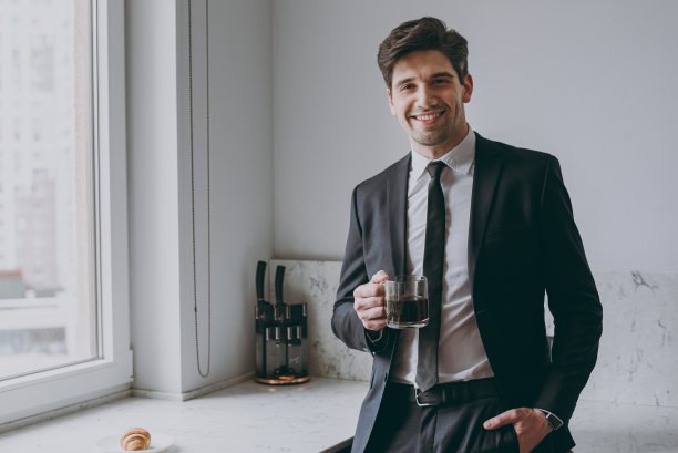 年轻，成功，自信，快乐的员工，商人，公司律师，二十多岁，穿着正式的黑色西装，衬衫，领带，喝咖啡，早餐，看相机，在轻厨房成就职业生活方式的概念图片下载