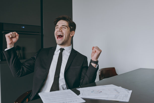 年轻的成功雇员，喜出眉外的商人公司律师穿着正式的黑色西装衬衫领带读纸文件做胜利者的姿势握拳坐在厨房的桌子旁。人们生活方式的概念图片下载