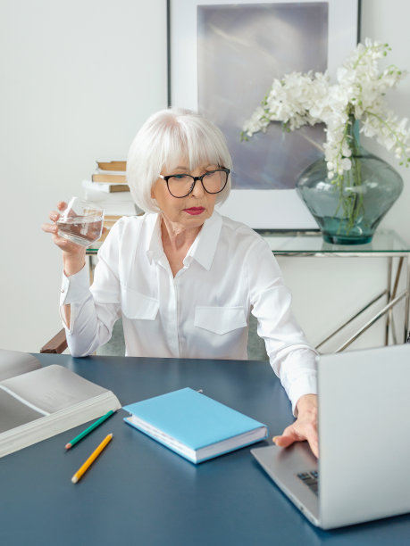 高级漂亮的灰色头发的女人在白色的衬衫在办公室里喝水。工作，资深人，水平衡，找到解决方案，体验理念图片下载