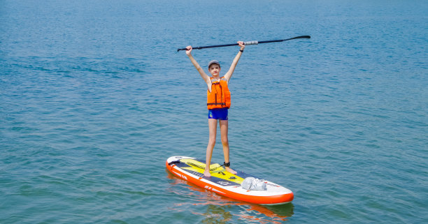 快乐微笑的男孩站在sup冲浪举起他的手，举着一个桨在他的头。成功，胜利，积极的生活理念。阳光明媚的一天，13-15岁的少年冲浪者在蓝色的波浪上冲浪图片下载
