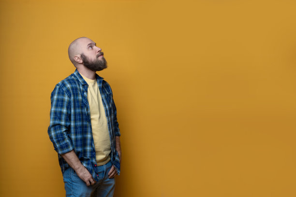 一名留着胡子的成年男性穿着格子衬衫的肖像，背景是黄色的。快乐的人看向右边孤立的黄色墙壁与副本空间。快乐成功自信的男人。图片下载