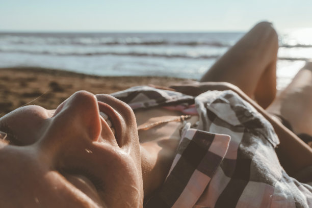 一个年轻快乐微笑的女人在泳衣和衬衫的特写躺在沙滩上对着大海的背景图片下载