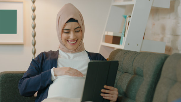 图为，在家中躺在沙发上用平板电脑与丈夫交谈的年轻穆斯林孕妇。图片下载