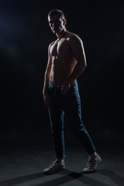 一个上身赤裸的肌肉男穿着牛仔裤在黑暗的摄影棚里摆姿势的剪影。图片下载