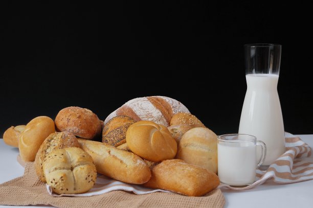 桌上的毛巾上放着新鲜出炉的面包制品——意大利脆皮面包、小圆面包、法棍面包、圆面包。旁边有一个罐子和一杯牛奶。这张照片是以黑色为背景拍摄的。图片下载