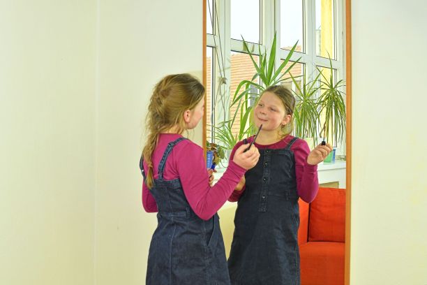 美丽时尚理念。孩子脸上有漂亮的笑容，在室内画嘴唇。小女孩拿着口红或睫毛膏站在房间的镜子前。孩子试着画嘴唇，用妈妈的化妆品化妆图片下载