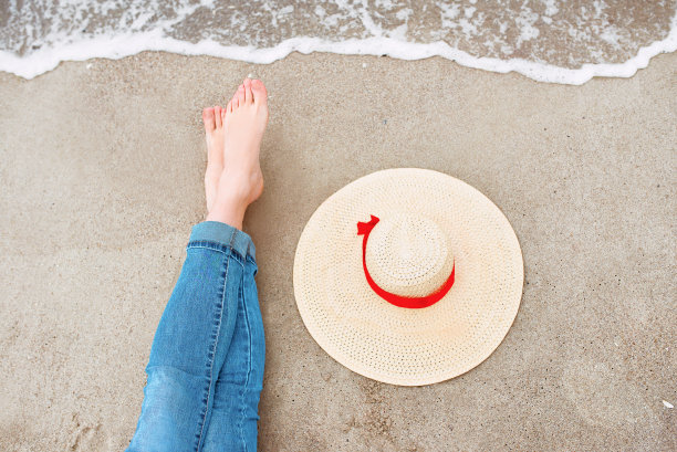 穿着牛仔裤的白人妇女的腿和脚坐在海边，旁边的沙滩上的帽子。旅行抽象照片图片下载