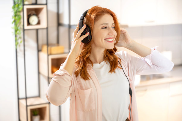 年轻漂亮的红发女人戴着耳机听音乐，而跳舞在家里。图片下载