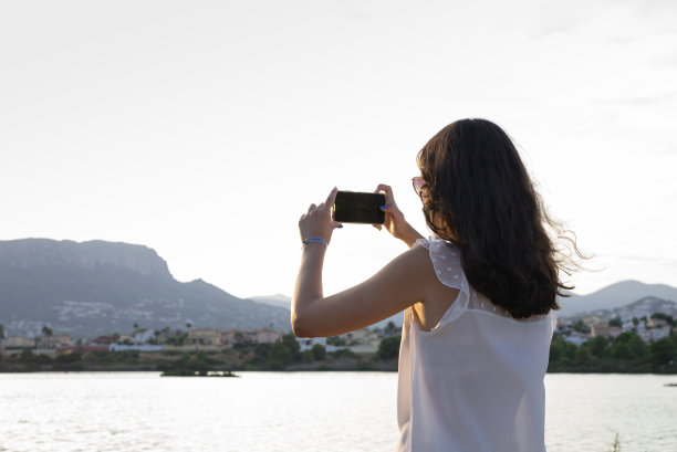 一个年轻的女孩，用她的手机拍摄日落。图片下载