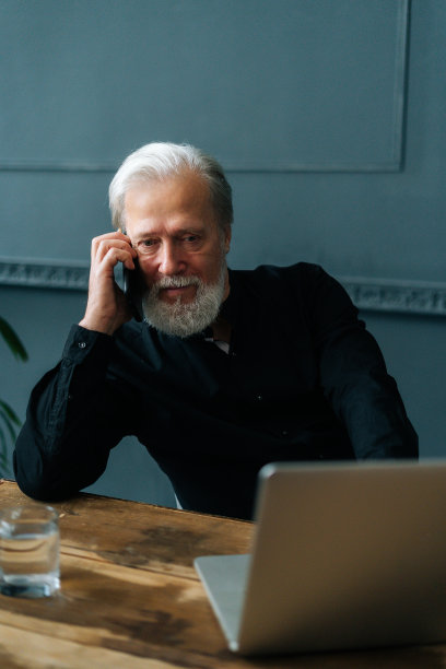 垂直肖像严肃，白发苍苍的成熟的商人，在家里的办公室里，坐在桌子旁用笔记本电脑打电话。图片下载