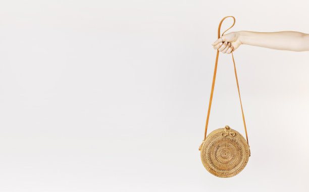 一个年轻女孩的手握着一个时尚的自然藤包，背景是一堵浅色的墙。来自巴厘岛的藤条手袋、环保袋。环保袋的概念，时尚的竹袋。本空间图片下载