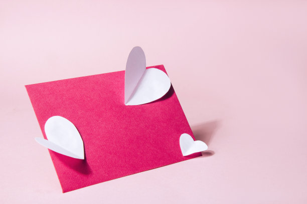 粉红色信封与白色的纸心在裸粉红色的背景。白色体积纸心图片下载