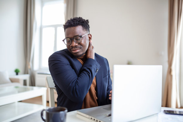 感觉筋疲力尽。沮丧的年轻英俊男子看起来筋疲力尽，坐在家里，在他的笔记本电脑上工作，同时按摩他的颈部，颈部疼痛图片下载