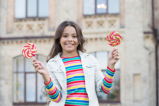 拿着棒棒糖的嬉皮士小孩。她友好的微笑。快乐的儿童的一天。小女孩长着一头卷发。春天节日的心情。小美心情积极。童年的幸福。丰富多彩和甜图片下载