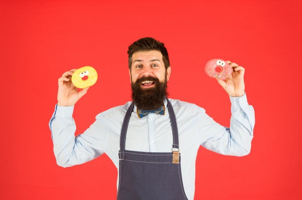 分享有趣的甜甜圈。大胡子男人拿着红色背景的环形甜甜圈。专业的面包师提供美味的甜甜圈。嬉皮士手里拿着油炸甜甜圈图片下载