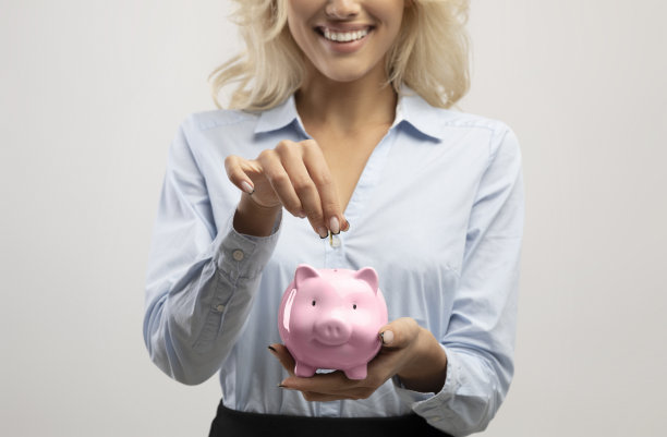 储蓄和投资观念。女商人把钱放在粉红色的存钱罐，浅灰色的工作室背景，作物图片下载