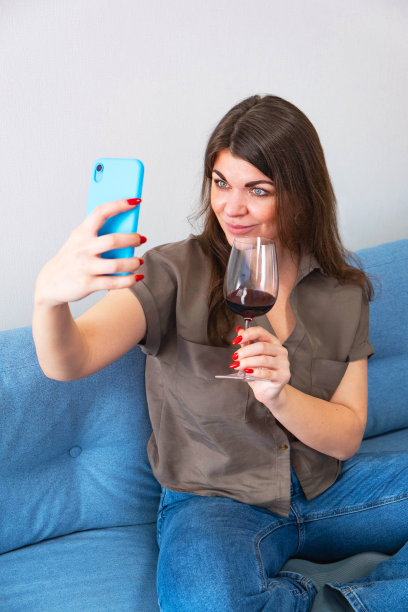 一名年轻女子在家里的沙发上拿着一杯红酒在手机上自拍。图片下载