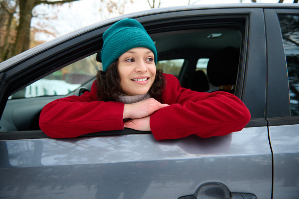 漂亮的女人，穿着鲜红的外套，戴着绿色的针织帽，坐在现代汽车的驾驶座上，行驶在白雪覆盖的道路上，透过打开的窗户，享受着外面凉爽的冬天图片下载