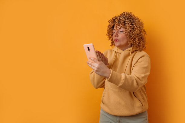 30多岁的女性博主看着智能手机上的相机，在黄色的背景下拍了一张自拍。人的概念图片下载