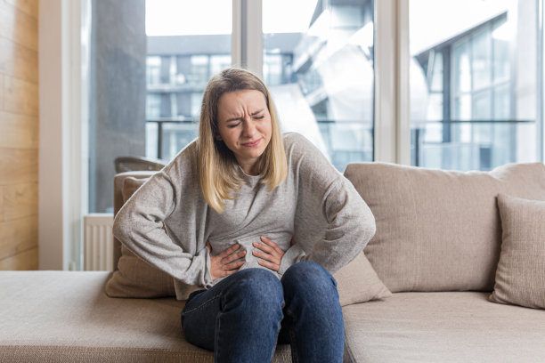 一个年轻的女人坐在家里的沙发上，抓着肚子疼，疼得大叫，她感觉疼，她不舒服，感觉很糟糕图片下载