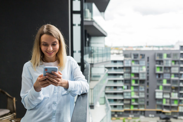 美丽的金发女子站在公寓楼的阳台上用手机图片下载