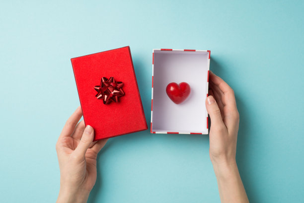 第一人称俯视图照片的情人节装饰女孩的手握小心脏在打开的礼品盒与红色的盖子和星弓在孤立的淡蓝色背景图片下载