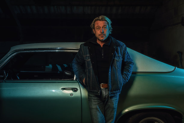 一个金发男子穿着牛仔夹克站在一辆美国经典肌肉车旁。图片下载