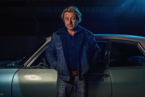 一个金发男子穿着牛仔夹克站在一辆美国经典肌肉车旁。图片下载