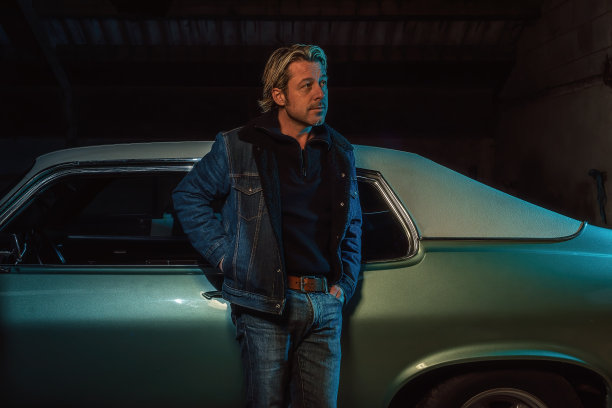 一个金发男子穿着牛仔夹克站在一辆美国经典肌肉车旁。图片下载