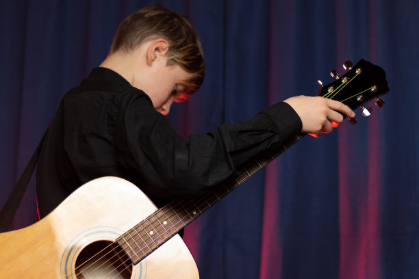 一个十几岁的男孩穿着黑色的衬衫，手里拿着吉他，紫色的背景和红色的灯光在舞台上。有选择性的重点。图片下载