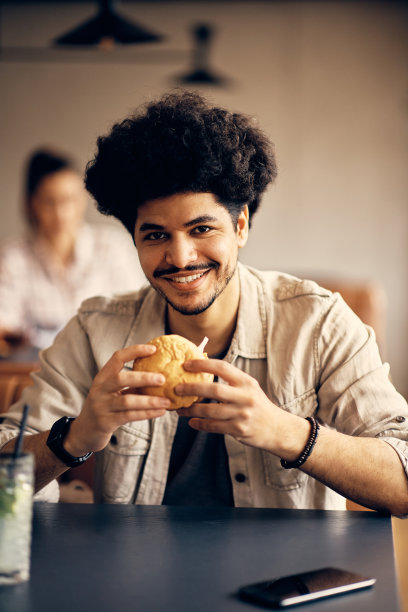 年轻快乐的穆斯林男子在自助餐厅吃汉堡，看着镜头。图片下载