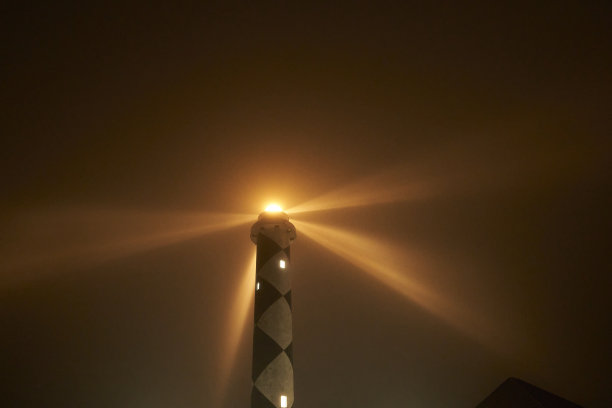 高大的黑白灯塔用光束照亮黑暗，透过夜晚的薄雾，为船只导航提供方向图片下载