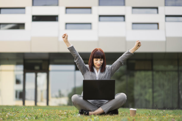 穿着西装的漂亮女人，坐在草地上用笔记本电脑，庆祝成功图片下载