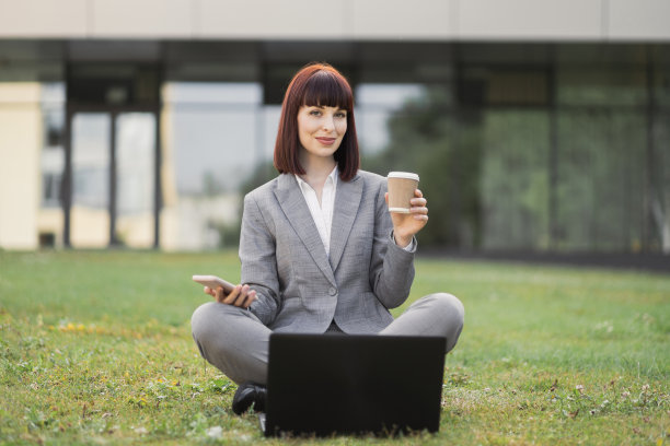 穿着灰色西装的年轻女商人坐在草地上，拿着笔记本电脑，送咖啡图片下载