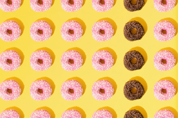 粉红色和巧克力色的环形甜甜圈。白色和棕色的小碎屑在黄色的粉彩背景上。模式。平的。图片下载