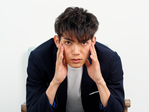 英俊的中国年轻人的肖像，在深蓝色的休闲西装坐在扶手椅和摆姿势的白墙背景。双手捂着脸，严肃地看着前面。图片下载