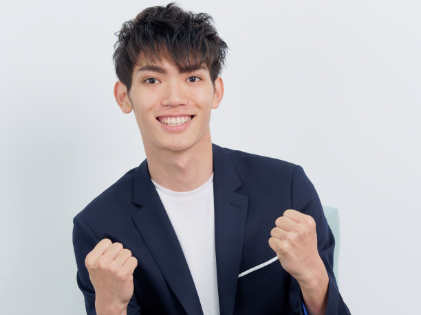 英俊的中国年轻人的肖像，在深蓝色的休闲西装坐在扶手椅和摆姿势的白墙背景。两个拳头举起来做胜利和力量的手势，正面看。图片下载