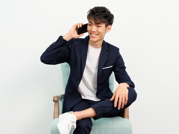 英俊的中国年轻人的肖像，在深蓝色的休闲西装坐在扶手椅和摆姿势的白墙背景。面带微笑，双腿交叉，用手机通话。图片下载