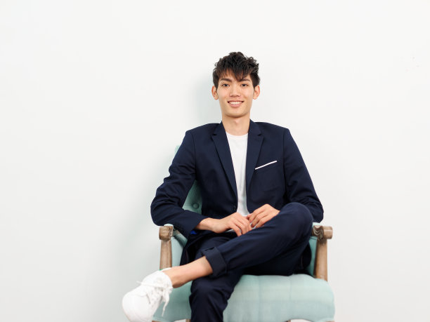 英俊的中国年轻人的肖像，在深蓝色的休闲西装坐在扶手椅和摆姿势的白墙背景。双腿交叉，对着镜头微笑，看起来很自信。图片下载