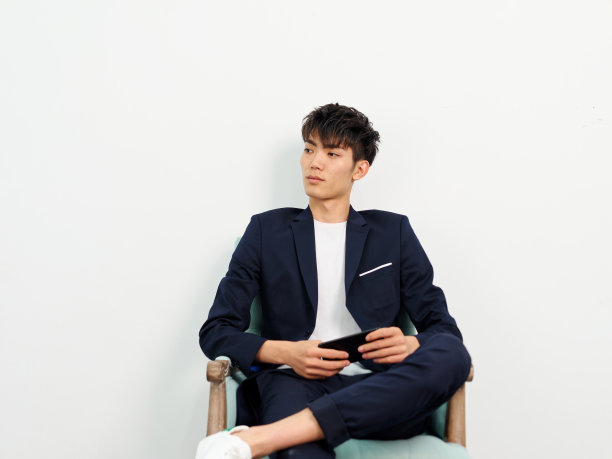 英俊的中国年轻人的肖像，在深蓝色的休闲西装坐在扶手椅和摆姿势的白墙背景。两腿交叉，手里拿着手机，目光移开，思考着。图片下载