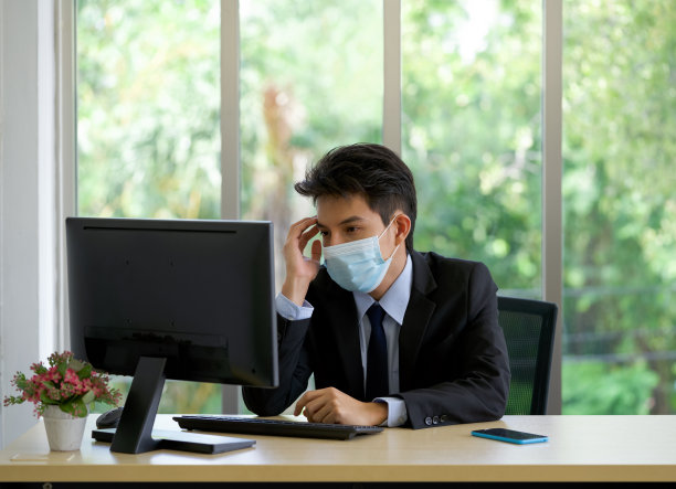 年轻的亚洲商人戴着口罩，在办公室工作时用一只手触摸太阳穴。图片下载