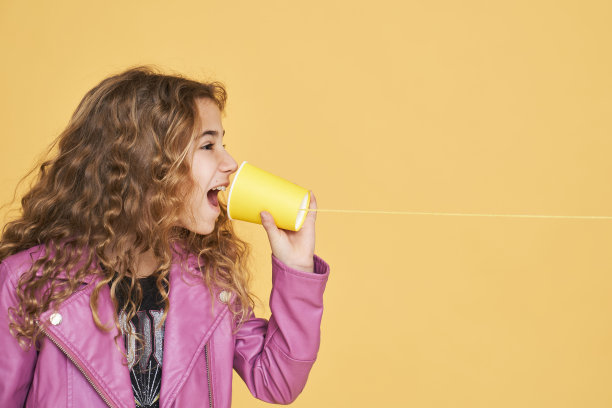 女孩透过一个纸杯说话，纸杯绑在一个黄色背景的线孤立。图片下载