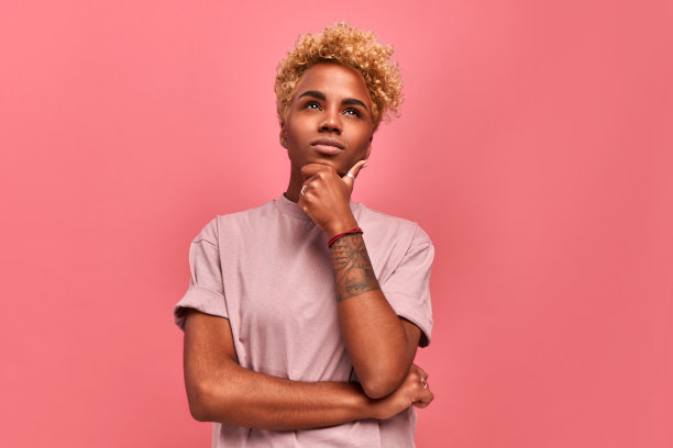 让我想想。深思熟虑的深色皮肤的女人，金色的头发，钱包嘴唇，手放在下巴下，计划着什么或做出选择，在心里做决定，站在粉红色的背景。怀疑的概念图片下载