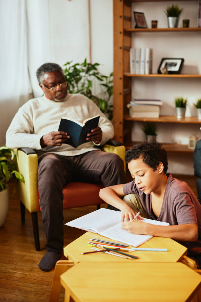 一位非洲爷爷坐在家里看书，陪着孙子。一个孙子正在做他的数学作业。代际和年龄差异图片下载